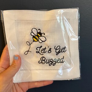 Könnte beinhalten: Weißes Cocktail-Servietten aus Leinen mit einer schwarzen, gestickten Biene und dem Schriftzug "Let's Get Buzzed". Die Serviette ist quadratisch und hat einen dekorativen, genähten Rand. Die Serviette ist in Klarsichtfolie verpackt.