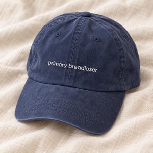 May include: A navy blue baseball cap with a six-panel construction and a curved brim. The front of the cap features the text 'primary breadloser' embroidered in white lowercase letters. The fabric has a slightly faded, washed-out appearance.