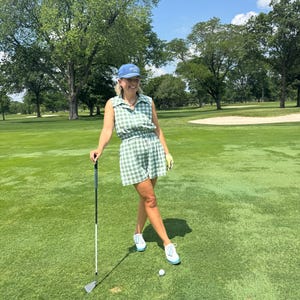 May include: A woman wearing a green and white checkered golf outfit, including a sleeveless top and shorts, and a blue cap. She is holding a golf club on a green golf course, with a golf ball nearby. The outfit is coordinated with white sneakers.
