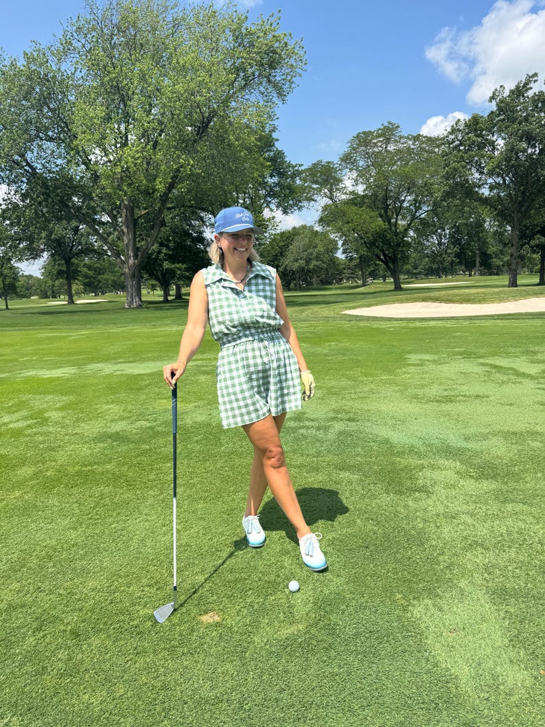 May include: A woman wearing a green and white checkered golf outfit, including a sleeveless top and shorts, and a blue cap. She is holding a golf club on a green golf course, with a golf ball nearby. The outfit is coordinated with white sneakers.
