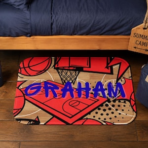 May include: A red and brown basketball-themed mat with the name "GRAHAM" in blue. The mat features basketballs, a hoop, and court lines. A blue water bottle and a navy blue bag are also visible.