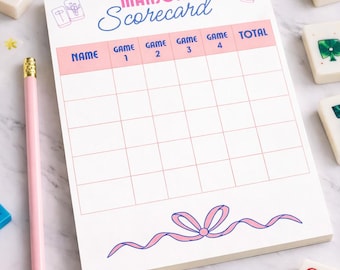 May include: A pink and white Mahjong Scorecard with a bow design, surrounded by Mahjong tiles and a pink pencil. The scorecard has spaces for names, game numbers, and totals.