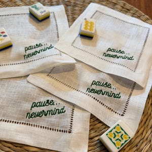 May include: Four square white linen napkins with hemstitched borders, each embroidered with the green text "pause... nevermind". Three small rectangular mahjong tiles with floral and numerical designs are placed on top of the napkins.