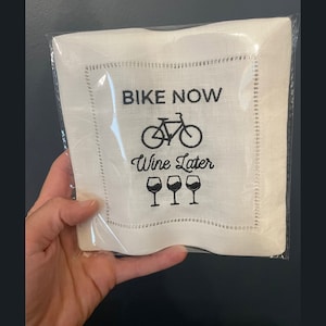 May include: White linen napkin with black embroidery that reads "Bike Now Wine Later" with a bicycle and four wine glasses.