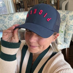 May include: A woman wearing a navy blue and black mesh trucker hat with the word "BIG" in red embroidered on the front. She is wearing a cream and green striped sweater.