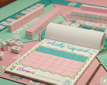 May include: A pink and teal Mahjong set with tiles and a score pad that reads "Mahj Squad" and "Winner". The set is on a pink and teal game board. The tiles are pink and white.