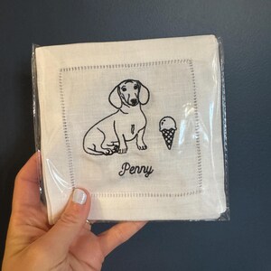 May include: White linen napkin with black embroidery of a dachshund dog and an ice cream cone. The napkin is personalised with the name "Penny".