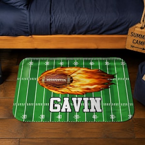 May include: A rectangular green football field-themed mat with white yard lines and the name "GAVIN" in bold white letters. A brown football with flames is in the center. The mat is on a wooden floor.