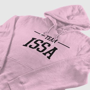Könnte beinhalten: Ein hellrosa Kapuzenpullover mit Kapuze und Vordertasche. Der Text "TEAM ISSA" ist in Schwarz auf die Brust gedruckt. Der Hoodie besteht aus einem weichen Material und wird auf weißem Hintergrund dargestellt.