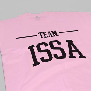 以下が含まれることがあります： 薄いピンク色のTシャツ。黒い太字で「TEAM ISSA」とプリントされています。「TEAM」は水平線の上に、そして「ISSA」は下に配置されています。柔らかい素材で作られています。