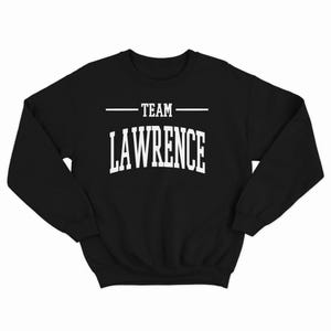 以下が含まれることがあります： 白い文字で「TEAM LAWRENCE」とアーチ型のフォントで書かれた黒いスウェットシャツです。
