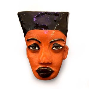 May include: A ceramic face sculpture with a dark brown, glossy headpiece and dark brown lips. The face is a warm orange color with dark eyes and eyebrows. The sculpture is a unique piece of art.