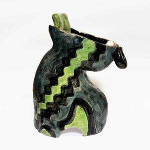 May include: A handcrafted ceramic vase with a unique, abstract design. The vase features a dark teal base with black and lime green zigzag and curved patterns. A small, green rectangular piece is on the top rim, and a black spout extends from the side.