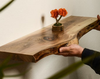 May include: A rustic wooden shelf with a live edge, made from natural wood with a rich brown colour and a unique grain pattern. A small orange bonsai tree sits on the shelf.