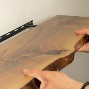 K&ouml;nnte beinhalten: Ein Holzregal mit einer lebenden Kante wird mit einer schwarzen Metallhalterung an einer Wand montiert. Das Regal besteht aus hellem braunem Holz mit dunkelbraunen Maserungen.