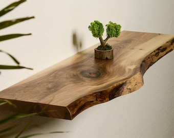 Puede incluir: Una estantería de madera natural con un borde vivo y un acabado rústico. La estantería está montada en una pared blanca y tiene una pequeña planta en maceta en la parte superior.