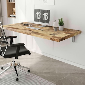 Puede incluir: Un escritorio de madera montado en la pared con un patrón de vetas de madera natural. El escritorio es rectangular y está sostenido por soportes plateados. Un monitor de computadora muestra los números "13 16". Una silla de oficina negra está frente al escritorio. Una pequeña planta y una botella de agua están en el escritorio.