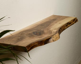 Puede incluir: Una estantería de madera natural con un borde vivo y un acabado rústico. La estantería está montada en una pared blanca.