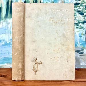 May include: A vintage hardback book titled "Through the Looking Glass" by Lewis Carroll. The cover is a light beige colour with a faded illustration of Alice holding a small object.