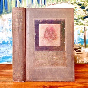 Pode incluir: Livro antigo de capa dura intitulado "Anne of Green Gables" de L.M. Montgomery. O livro tem uma capa marrom desbotada com um retrato de uma mulher. A lombada também é marrom com o título e o nome do autor. O livro está em uma superfície de madeira.