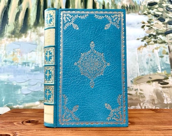 Puede incluir: Un libro de tapa dura azul turquesa con detalles dorados y un diseño decorativo en la portada. El libro está sobre una superficie de madera.
