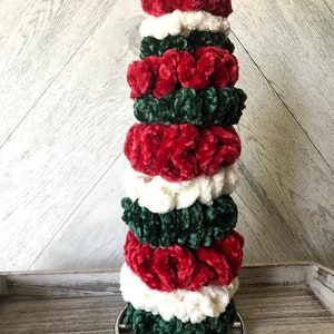 May include: A stack of eight scrunchies in red, white, and green velvet. The scrunchies are arranged in a tower on a silver stand.