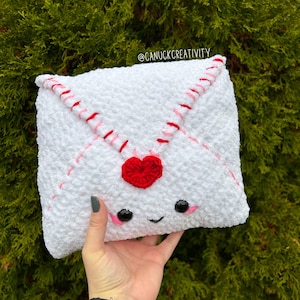 May include: White crocheted envelope pillow with red and pink stitching, a red heart, and a smiling face with black eyes. The pillow is being held by a hand.