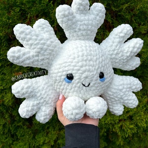 Puede incluir: Juguete de peluche de copo de nieve blanco tejido a crochet con ojos azules y una boca sonriente. El juguete está siendo sostenido por una mano que lleva una sudadera negra. El fondo es un arbusto verde.
