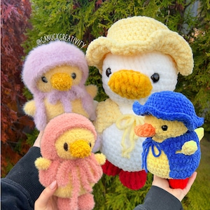 May include: Four crocheted duck plushies in different colors and styles. The ducks are wearing hats and have yarn eyes. The ducks are sitting on a green surface. @ganuckcreativity