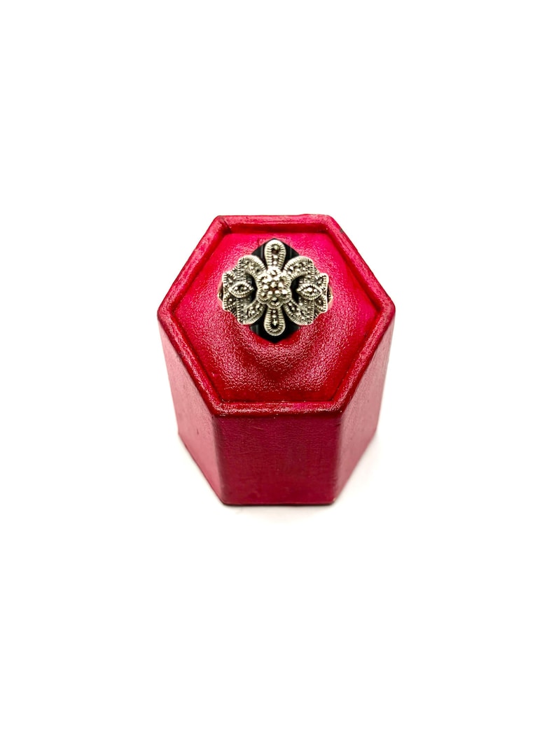 A silver ring with a black onyx center and marcasite floral design, displayed on a red hexagonal ring stand, viewed from the top.