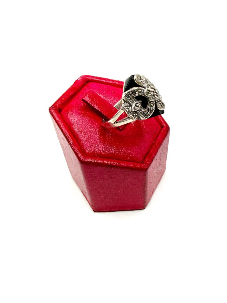 A silver ring with a black onyx center and marcasite floral design, displayed on a red hexagonal ring stand, viewed from the side.