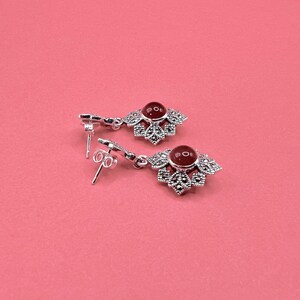 A pair of silver earrings with red carnelian centers and marcasite accents, lying on a pink surface.