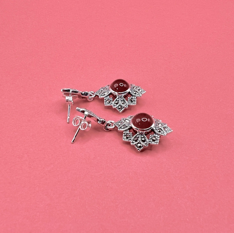 A pair of silver earrings with red carnelian centers and marcasite accents, lying on a pink surface.