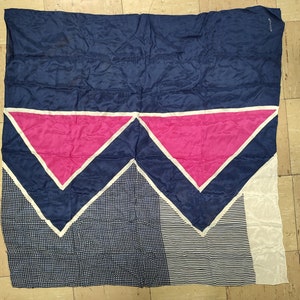 May include: A large, rectangular scarf with a geometric design. The top portion is navy blue, followed by pink triangles outlined in white. The bottom features a white polka dot pattern and white stripes. The scarf is made of a silky material.