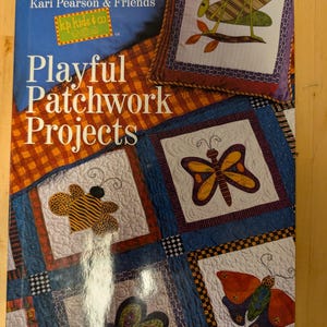 May include: Book cover featuring a patchwork quilt with a variety of colorful designs, including butterflies, a bee, and a grasshopper. The title of the book is "Playful Patchwork Projects" by Kari Pearson & Friends.