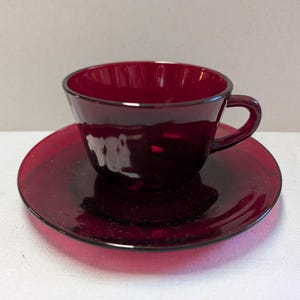 May include: A deep ruby red glass teacup and matching saucer set. The teacup features a smooth, rounded bowl shape with a small, curved handle on the side. The saucer is circular with a slightly raised center to hold the cup securely in place.
