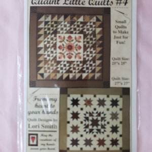 May include: A package featuring two quilt patterns. The top quilt design has a central floral motif and geometric shapes in brown, tan, and cream. The bottom quilt design showcases a star pattern in similar colors. Text includes "Quaint Little Quilts #4" and "From my heart to your hands."