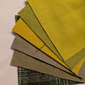 May include: A fan of fabric swatches in shades of green, yellow, and gray. The samples include solid colors and a plaid pattern, showcasing various textures and hues. The fabrics are arranged on a white surface, providing a clean backdrop for the color palette.