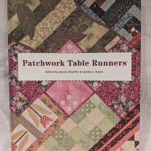 May include: A book titled "Patchwork Table Runners" with a colorful patchwork design on the cover. The cover features various fabric patterns in shades of pink, green, and brown, with white buttons and the text "Edited by Jeanne Stauffer & Sandra L. Hatch".