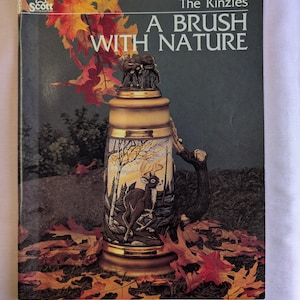 May include: A book titled "The Kinzies A Brush With Nature" featuring a ceramic stein with a deer and autumn leaves painted on it.