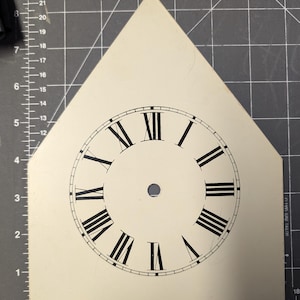 May include: A white clock face with black Roman numeral markings and a black outline. The clock face is mounted on a white triangular piece of cardboard.
