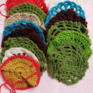 May include: A collection of handmade crochet coasters in various colors, including red, green, white, and brown. Each coaster features a circular design with a scalloped edge and a loop for hanging. The coasters are arranged on a pink surface.