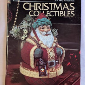 May include: A book titled "Christmas Collectibles" by Duncan Designers featuring a hand-painted Santa Claus figurine holding a lantern. The Santa Claus is wearing a red and white suit with a green hat and a white beard.