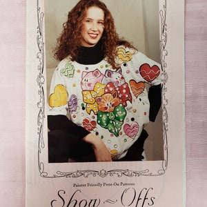 May include: A woman wearing a white sweatshirt with a colorful cat and heart design. The sweatshirt is part of a book cover titled "Show Offs" with the text "Painter Friendly Press-On Patterns" and "designs by devonia". The image is framed.