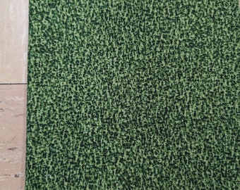 May include: Close-up of a folded fabric with a textured pattern. The fabric is primarily green with a speckled design in a darker shade of green, creating a mottled appearance. The material appears to be soft and pliable.