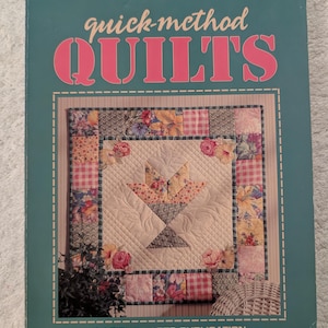 May include: A teal-colored book cover with the title "quick-method QUILTS" in pink lettering. The cover features a quilt design with floral and checkered patterns, centered around a basket motif. The text "A LEISURE ARTS PUBLICATION" is at the bottom.