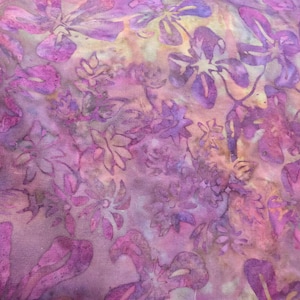 May include: A piece of fabric featuring a batik-style floral pattern in shades of purple, magenta, and soft yellow. The design consists of repeating flower and leaf motifs layered over a mottled, textured background with visible color gradients.