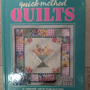 May include: A teal hardcover book titled quick-method QUILTS, A LEISURE ARTS PUBLICATION. The cover features a central image of a quilted basket design surrounded by a border of floral and checkered fabric squares in pink, green, and yellow.