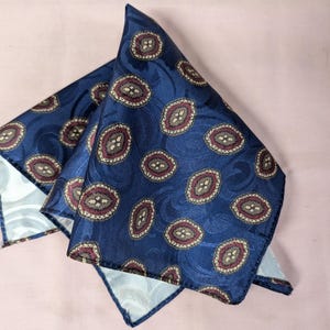 May include: A blue silk pocket square with a paisley pattern in shades of red, brown, and white.