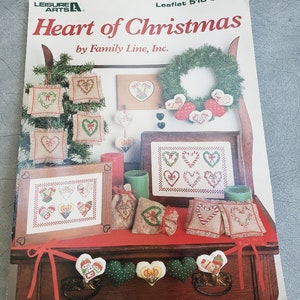 May include: A craft leaflet titled "Heart of Christmas" by Family Line, Inc. The cover features various Christmas-themed cross-stitch projects, including hearts, wreaths, and small framed pieces. The color palette includes red, green, and beige.